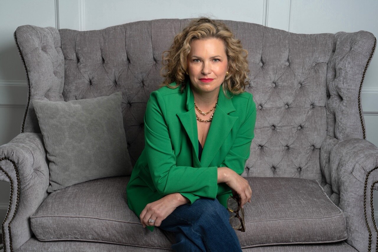 Julia Hoffman in a green blazer and jeans, sitting on a gray tufted sofa and smiling at the camera